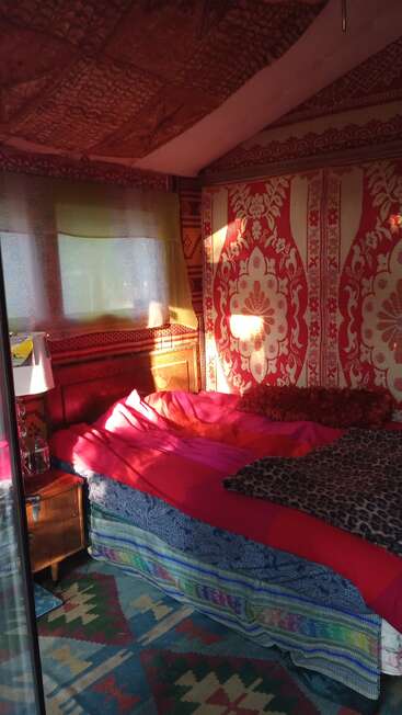 Ein gemütliches, farbenfrohes Schlafzimmer mit lebhaft gemusterten Teppichen, Decken und Wandteppichen. Warmes Sonnenlicht fällt durch ein Fenster und wirft Schatten auf ein ungemachtes Bett und einen Nachttisch.