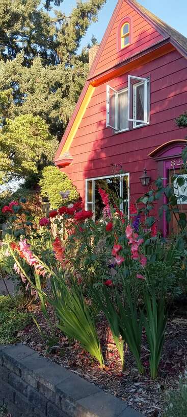 Ein charmantes rotes Haus mit offenem Fenster ist von üppigem Grün und blühenden Blumen umgeben und sonnt sich im warmen, goldenen Sonnenlicht unter hohen Bäumen.