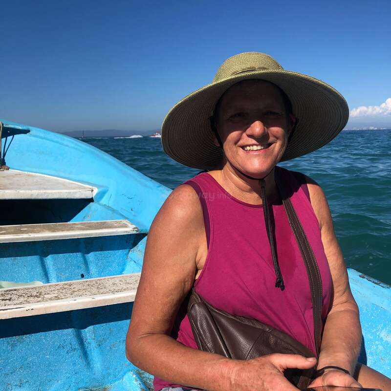 Eine lächelnde Frau, die einen breitkrempigen Hut und ein ärmelloses Hemd trägt, sitzt auf einem blauen Boot und genießt einen sonnigen Tag auf dem Wasser unter einem klaren Himmel.