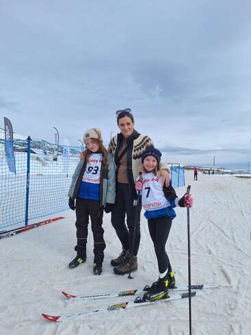 A woman stands between two smiling children in ski gear on a snowy slope, posing for a photo. Blue nets and flags are visible in the background.