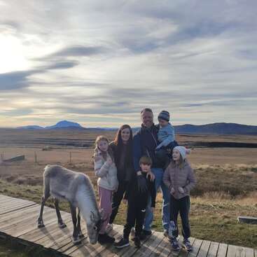 Eine sechsköpfige Familie posiert auf einem Holzweg in einem weiten, offenen Feld mit Bergen im Hintergrund. Ein junges Rentier grast in der Nähe.