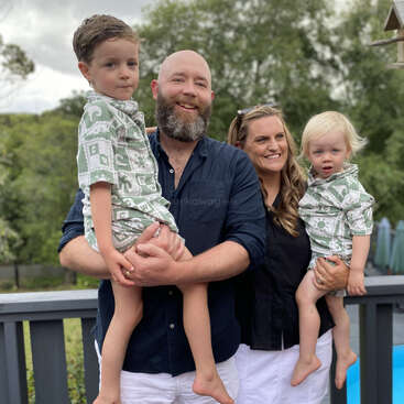 Eine glückliche vierköpfige Familie posiert im Freien, die Eltern halten ihre beiden kleinen Jungen im Arm. Die Jungen tragen passende Outfits, alle lächeln und sind von Grün umgeben.