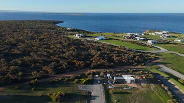 A coastal landscape with a dense forest, scattered houses, and grassy fields. Roads wind through the area, leading to the calm blue ocean in the background.
