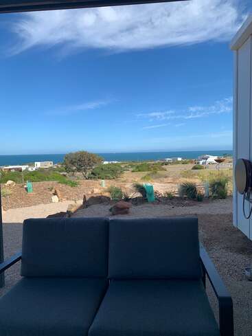 A cozy outdoor sofa overlooks a serene, coastal landscape with scattered houses, green shrubbery, blue ocean, and a vast, beautiful sky filled with wispy clouds.