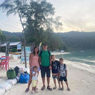 A imagem mostra uma família de cinco pessoas posando em uma praia de areia, com um corpo de água sereno e uma vegetação exuberante ao fundo, evocando uma sensação de tranquilidade.
