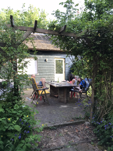 The image depicts a serene outdoor setting, featuring a wooden table and chairs on a patio, surrounded by lush greenery and a charming house with a thatched roof in the background.