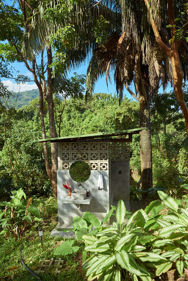 Un lavabo de exterior con un espejo circular y una pared de azulejos se alza en medio de una exuberante vegetación tropical. La luz del sol se filtra entre los altos árboles, creando una serena atmósfera selvática.