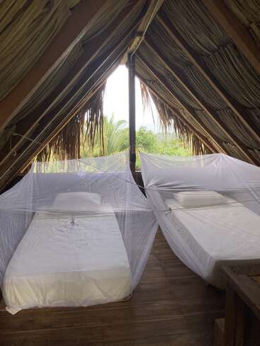 Esta rústica habitación abuhardillada cuenta con dos camas individuales con sábanas blancas, cada una cubierta por mosquiteras. El techo de palma y el exuberante paisaje verde del exterior crean un ambiente tropical.