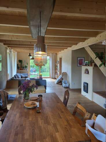 Dieser gemütliche, offene Wohnbereich besticht durch Holzbalken, einen rustikalen Esstisch mit Blumen, ein modernes Sofa, Zimmerpflanzen, viel Tageslicht und einen Blick auf den Garten.