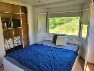 Ein gemütliches Schlafzimmer mit einem großen Fenster, das einen malerischen Ausblick bietet. Das Bett ist mit blauer Bettwäsche und Kissen bezogen, daneben steht ein Bücherregal aus Holz mit Büchern und Aufbewahrungsboxen.