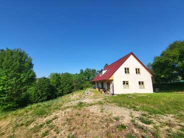 Ein charmantes Haus mit rotem Dach steht auf einem grasbewachsenen Hügel, umgeben von üppig grünen Bäumen unter einem klaren, strahlend blauen Himmel, und strahlt Ruhe aus.