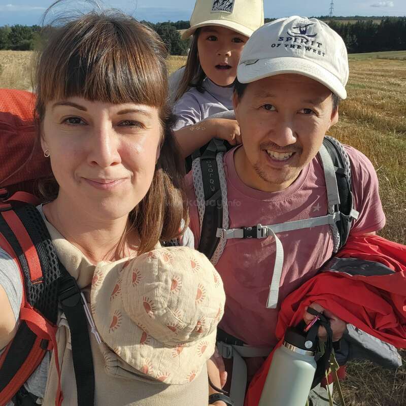 Eine vierköpfige Familie in Wanderkleidung lächelt auf einer sonnigen Wiese. Zwei Kinder werden von ihren Eltern in Rucksäcken getragen, die glücklich und abenteuerlustig aussehen.
