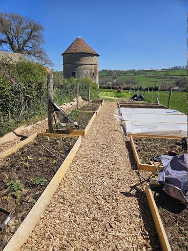 Un camino de virutas de madera bordea unos arriates elevados bajo un cielo azul despejado, con un edificio redondo de piedra y verdes campos de fondo.
