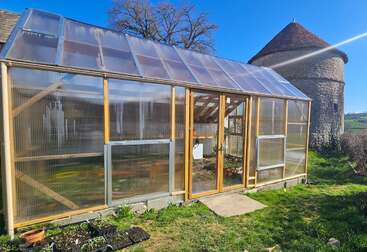 Ein sonniger Tag in einem Garten auf dem Land mit einem modernen Gewächshaus mit klaren Scheiben, Holzrahmen, grünen Pflanzen im Inneren und einem alten Steingebäude in der Nähe.