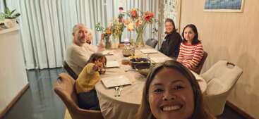 Five people are gathered around a dining table, smiling for a group selfie. The table is set for a meal, with flowers and dishes present. Cozy ambiance.