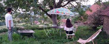 A man with a wheelbarrow stands near a woman sitting at a table under a colorful umbrella in a peaceful, grassy garden with rural houses.