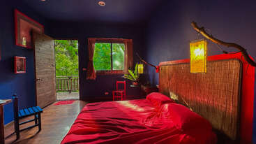 This bedroom features deep blue walls, a vibrant red bed, rustic wood accents, large green window views, and warm yellow lighting, creating a cozy, artistic atmosphere.