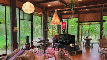 A cozy, sunlit music room with large windows, lush green views, musical instruments, cozy couches, artistic lighting, a piano, and eclectic decor creating a creative atmosphere.