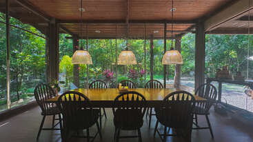 A wooden dining table with eight chairs sits under pendant lights. Large windows reveal a lush green garden, blending indoor comfort with serene outdoor nature.