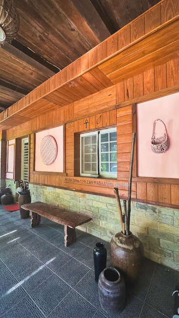 A rustic wooden porch features a bench, large pottery jars, woven baskets on the walls, stone accents, and a window, creating a cozy, traditional atmosphere.