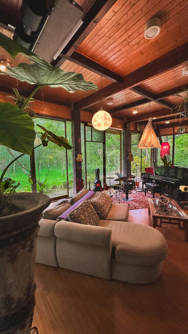 This cozy living room features large windows, wooden ceilings, indoor plants, eclectic hanging lamps, a beige sectional sofa, musical instruments, and lush greenery outside. Warm, artistic atmosphere.
