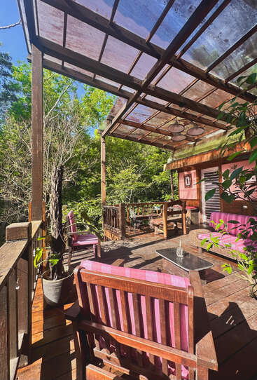 A wooden deck with pink cushioned chairs sits under a transparent roof, surrounded by lush greenery and sunlight, creating a cozy, inviting outdoor retreat in nature.