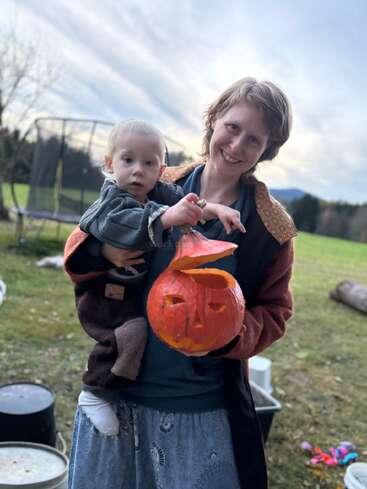 Una mujer con un niño pequeño y una calabaza tallada al aire libre, ambos sonriendo. El fondo muestra hierba, árboles, una cama elástica y un cielo nublado al atardecer.