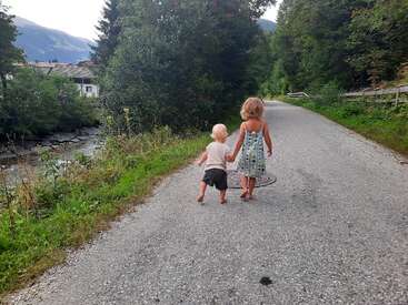 Dos niños caminan de la mano por una tranquila carretera rural bordeada de árboles, rodeados de exuberante vegetación y montañas, evocando una sensación de inocencia y aventura.
