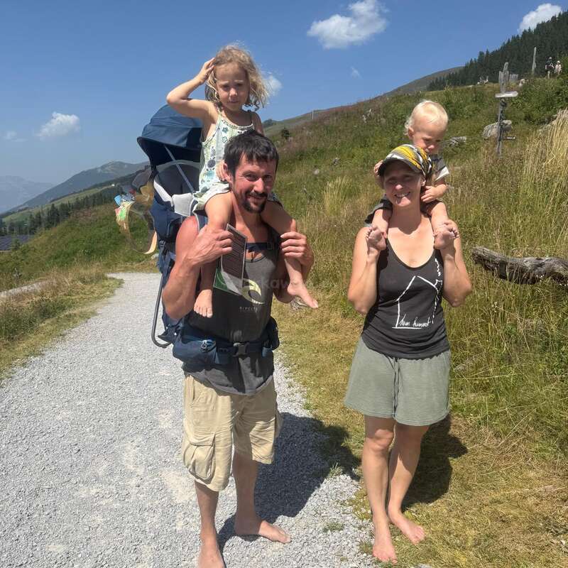 Una familia de cuatro miembros disfruta de un soleado día de excursión al aire libre, con los niños subidos a los hombros de los padres, sonrientes y descalzos por un camino de grava con pintorescas colinas.