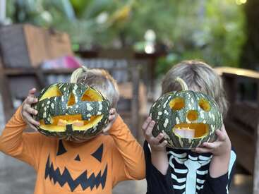 Duas crianças vestidas com roupas temáticas de Halloween seguram abóboras verdes esculpidas com rostos na frente de suas cabeças, fingindo ter cabeças de jack-o'-lantern ao ar livre.