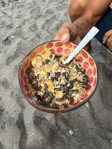 Une personne tenant un bol coloré rempli de muesli, de graines et de fruits secs, assise sur un sol sablonneux à la plage, prête à manger avec une cuillère.