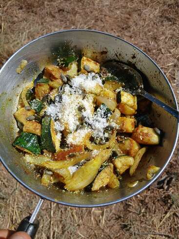 En un día soleado, se pone al aire libre una sartén metálica llena de verduras salteadas, incluidos calabacines y cebollas, cubiertas con queso rallado y semillas de calabaza.