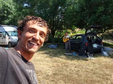 Un hombre sonríe para un selfie al aire libre, con una mujer tocando la guitarra cerca de un coche y una furgoneta. En el fondo soleado se ven árboles y material de acampada.