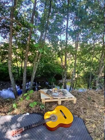 Una guitarra acústica amarilla descansa sobre una manta junto a un pequeño arroyo. Cerca, un palé de madera sostiene piedras y una flauta. La luz del sol se filtra entre los árboles.