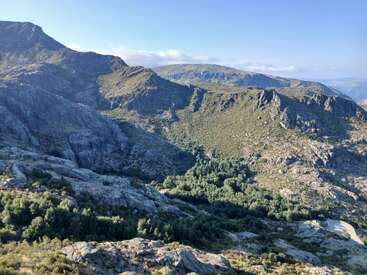 Bajo un cielo azul despejado se extienden ondulantes colinas verdes y escarpadas montañas rocosas, salpicadas de densos bosques, que crean un paisaje natural sereno y virgen.