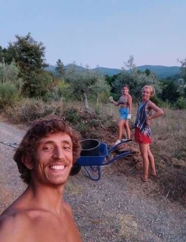 Three people outdoors, one taking a selfie with a big smile, the others standing behind near a blue wheelbarrow, surrounded by trees and hills at sunset.