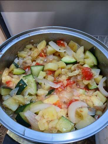 A metal pot filled with a colorful vegetable stew containing chopped zucchini, potatoes, onions, and tomatoes, cooked together into a hearty, rustic, homemade meal.