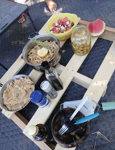 This image shows a rustic outdoor meal setup on a wooden pallet, featuring pasta, salad, jarred olives, seasonings, drinks, and utensils, with bare feet visible.