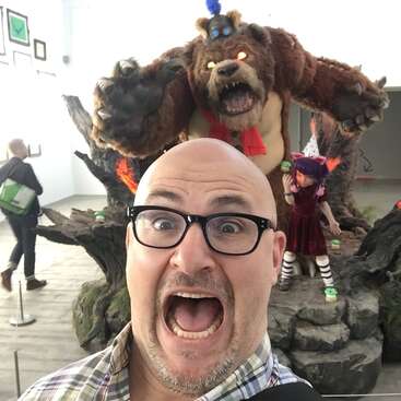 Das Bild zeigt einen Mann, der ein Selfie vor einem großen ausgestopften Bären und einem Mädchen in einem roten Kleid macht, vor dem Hintergrund eines Museums oder einer Kunstgalerie.