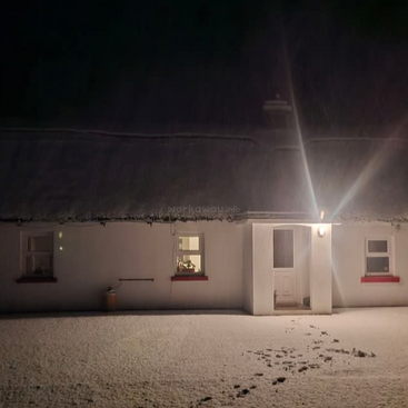 A cozy white cottage stands under a snowy night sky, warmly lit at the entrance. Footprints in the fresh snow lead to the inviting door.