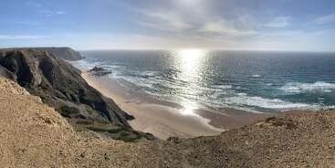 Una impresionante vista de la costa con acantilados escarpados, una playa de arena dorada y suaves olas que rompen en la orilla bajo un sol radiante que se refleja en el inmenso océano azul.