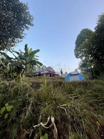 The image depicts a serene scene of a blue and white tent nestled among lush greenery, with a red house and trees visible in the background.