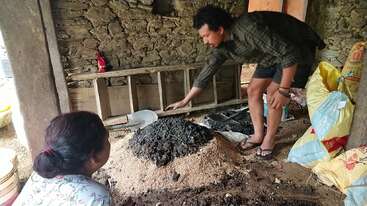 The image depicts a man and woman in a rustic stone building, surrounded by various materials, with the man bent over a pile of dirt and the woman sitting nearby.