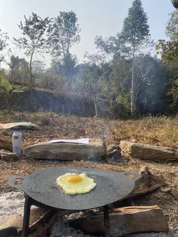 The image depicts a sunny outdoor scene with a frying pan containing a fried egg over a fire pit, surrounded by logs and trees in the background.