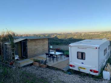 A white caravan and wooden cabin sit on a small wooden deck with outdoor furniture, overlooking a scenic green valley under a clear blue sky.