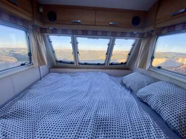 A cozy bed with patterned bedding inside a camper van, surrounded by large windows offering a stunning panoramic view of a scenic, sunlit landscape outside.
