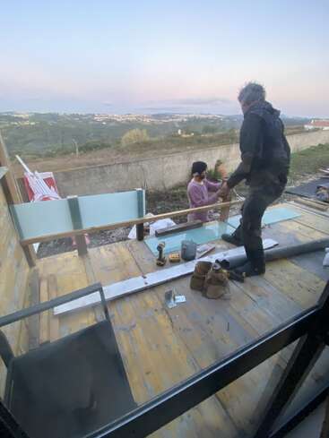Two people are building or repairing a wooden deck outdoors with tools and construction materials around them. There is a scenic landscape view in the background.