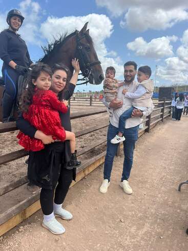 Una familia feliz posa junto a un caballo marrón en un rancho. Dos adultos sostienen a tres niños, mientras otra mujer monta el caballo. El cielo está nublado.