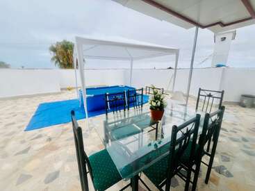 Esta imagen muestra un amplio patio exterior con una mesa de comedor de cristal, sillas verdes, un centro de mesa con plantas en macetas, un toldo y una piscina azul sobre el suelo.