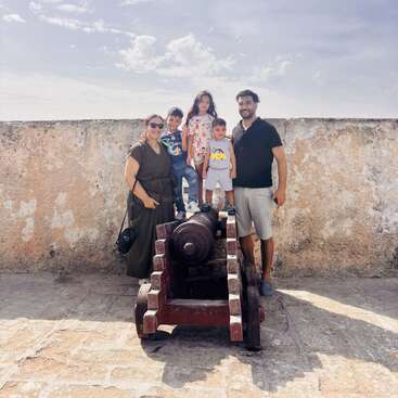 A family of five poses happily around an old cannon. The children stand on it while parents smile beside them. Bright, sunny day with blue skies.